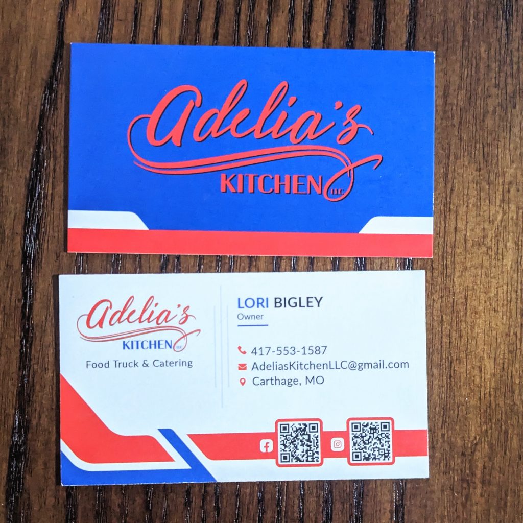 Business Card is on a matte finish . Primarily blue with Adelia's Kitchen in red on the front and the bottom has white block on both side and red blocking around bottom. The back is black and white with contact information as well as location.