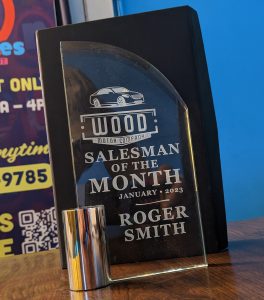 Glass rounded edge at top award with the words "Wood Motor Company Salesman of the Month January 2023 Roger Smith" all etched on it from top to bottom